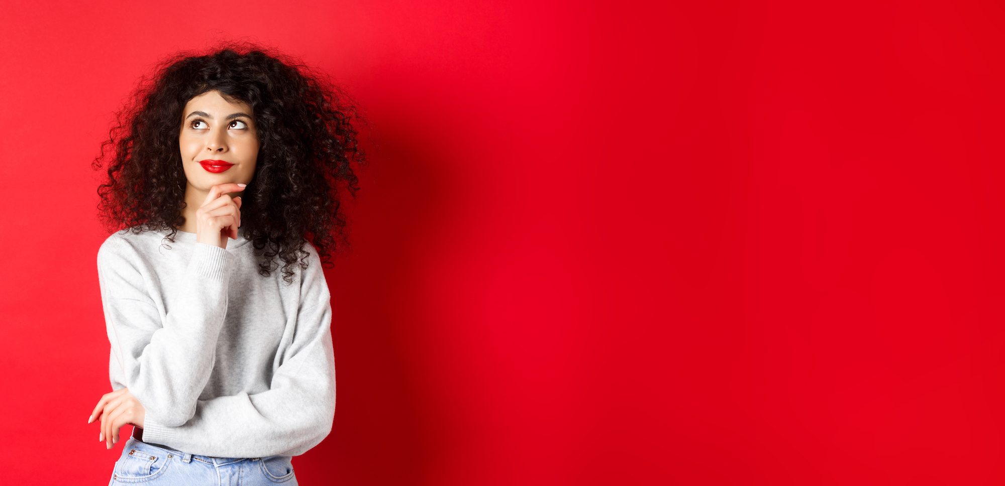 Pensive stylish woman with curly hair, red lips, looking thoughtful at upper left corner logo and smiling, standing against studio background.